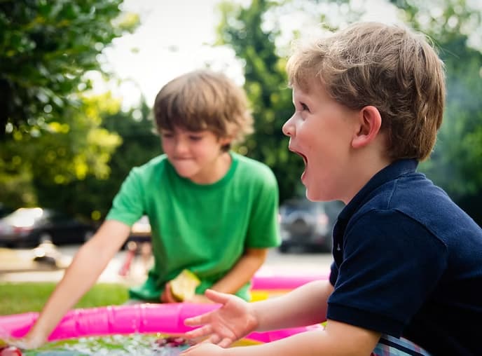 Two young boys play outside by a small pink pool. One in a green shirt leans forward while the other, in a navy shirt, smiles and gestures excitedly. Trees and blurred cars—plus a Viking HVAC top-rated Phoenix HVAC contractor van—are visible in the background.