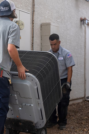 Two workers from Viking HVAC, a top-rated Phoenix HVAC contractor, move a large air conditioning unit on a dolly outside a building. One wears a cap while the other, in uniform with a U.S. flag patch, guides the unit near the wall.